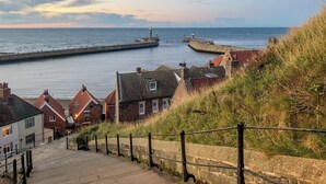 A coastal town with a stone staircase leading down to the beach, houses with red roofs, and a lighthouse in the distance.