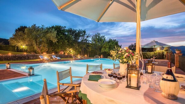 Un área de piscina al aire libre con sillones de descanso, una mesa de comedor con platos, vasos y velas, rodeada de árboles y un cielo despejado.