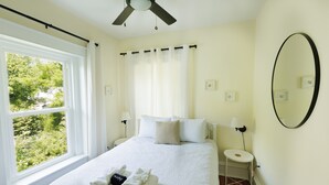 A neatly made bed with a white comforter, two pillows, and a folded towel on the bed. There is a window with white curtains, a ceiling fan, and a round mirror on the wall. - Serene Beach Haven in Cape Charles (Cape Charles)