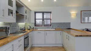 Een moderne keuken met witte kastjes, een houten aanrecht, een inbouwoven, een spoelbak en een raam met jaloezieën.