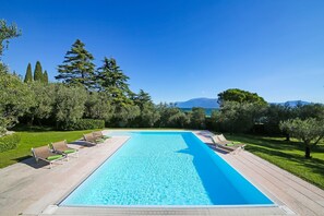 Una piscina rettangolare con un prendisole di legno e lettini, circondata da alberi e un cielo sereno.