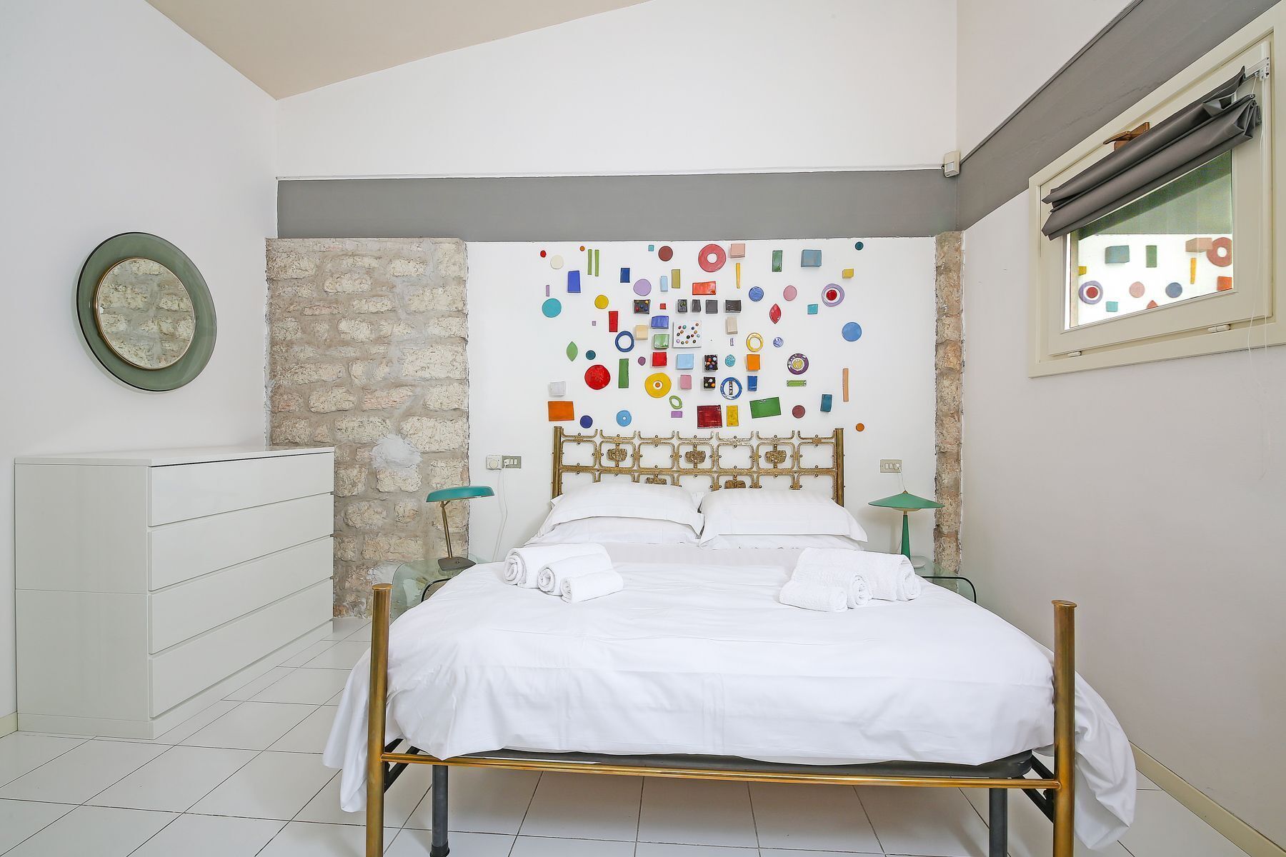 A bedroom with a bed, a dresser, a wall with colorful geometric shapes, and a window with a view.