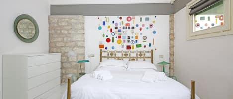 A bedroom with a bed, a dresser, a wall with colorful geometric shapes, and a window with a view.