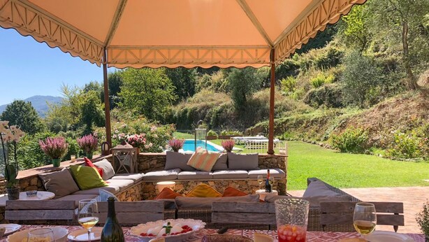Ett utomhusområde med plats för måltider, där ett bord är dukat, och som omges av en takförsedd sittplats med utsikt över en pool och kullar.