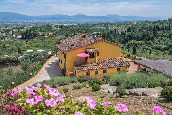 A yellow villa with a tiled roof, surrounded by lush greenery and a winding road.