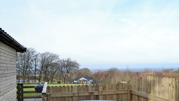 A hot tub in a wooden enclosure with a view of a residential area and trees in the background. - Kestrel Cabin - UK1246 (Brocklebank, near Caldbeck and Uldale)