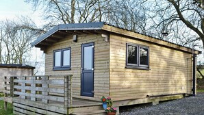 A wooden cabin with a dark door, two windows, and a small porch with a potted plant. - Kestrel Cabin - UK1246 (Brocklebank, near Caldbeck and Uldale)