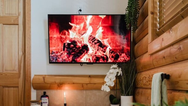 A spa room with a large bathtub, a mounted TV displaying a fire, and a wooden wall. - Jacuzzis & Family Kitchen & Entertainment Area (North Utica)