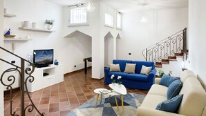 A living room with a blue sofa, beige sofa, and a TV on a white stand. There are shelves with decorative items and a staircase with a wrought iron railing. - Villa Minerva - Three Bedroom Villa, Sleeps 10 (Sant'Agnello)