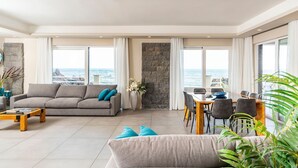 A spacious living area with a sofa, coffee table, and dining table, large windows with ocean views, and a stone accent wall. - Villa Sul Mare - Eight Bedroom Villa, Sleeps 16 (Acireale)