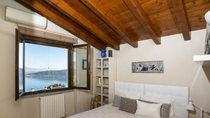 A bedroom with a large window, wooden ceiling beams, a bed with patterned bedding, a bedside table, and a shelf with decorative items. - Villa Meina - Four Bedroom Villa, Sleeps 8 (Meina)