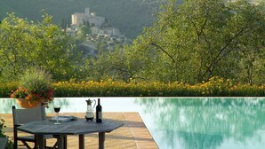 A poolside table with a wine bottle and glass, surrounded by greenery and a view of a castle in the distance.