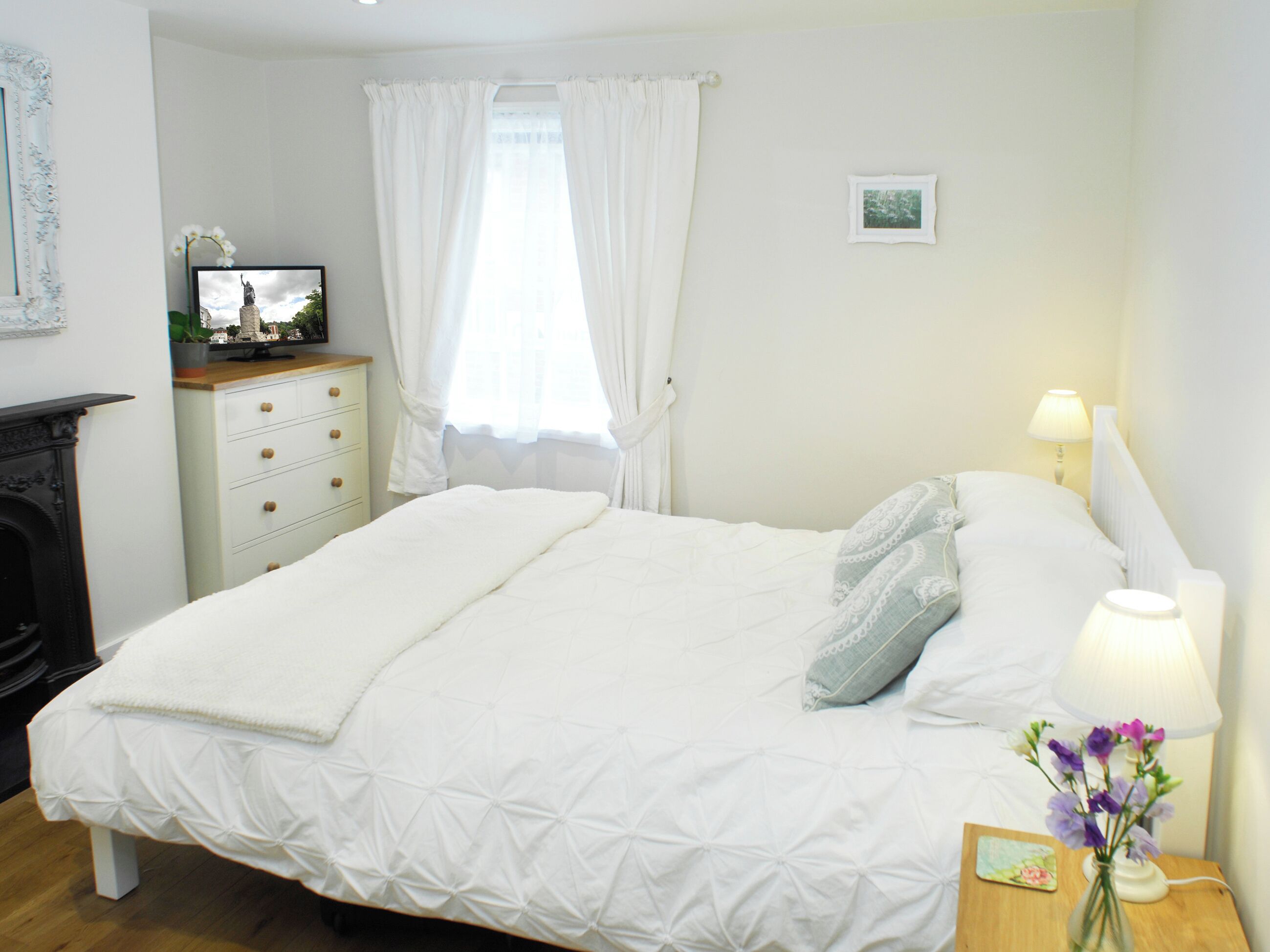 Un lit bien fait, avec une couette blanche et des oreillers, une table de chevet avec une lampe et un vase de fleurs, une commode avec des tiroirs, un tableau encadré au mur et une fenêtre avec des rideaux.