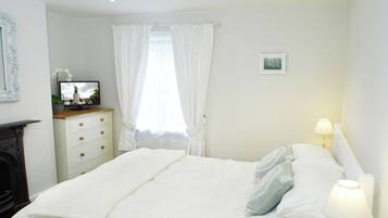 Un lit bien fait, avec une couette blanche et des oreillers, une table de chevet avec une lampe et un vase de fleurs, une commode avec des tiroirs, un tableau encadré au mur et une fenêtre avec des rideaux.