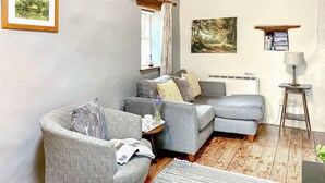 A cozy living room with a grey sofa, a small side table, a bookshelf, and a framed landscape painting. - Rebecca Puddle Duck Cottage (Bassenthwaite)