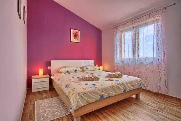 A bedroom with a wooden bed, a nightstand, a bedside lamp, a patterned rug, and a window with curtains.