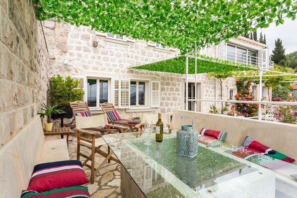 Un bâtiment en pierre avec une table en verre, des sièges à l’extérieur et une pergola couverte de plantes grimpantes.