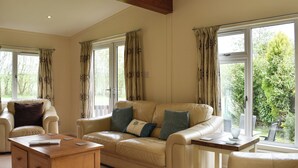 A spacious living room with a beige leather sofa, a wooden coffee table, and a dining table with chairs. Large windows provide views of greenery outside. - 3 bedroom accommodation in Lanreath, near Looe (Lanreath, near Looe)