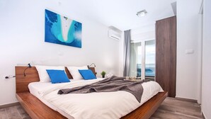 Ein modernes Schlafzimmer mit einem hölzernen Bett, weißer Bettwäsche, einem blauen Kunstwerk und Meerblick durch eine Glastür.