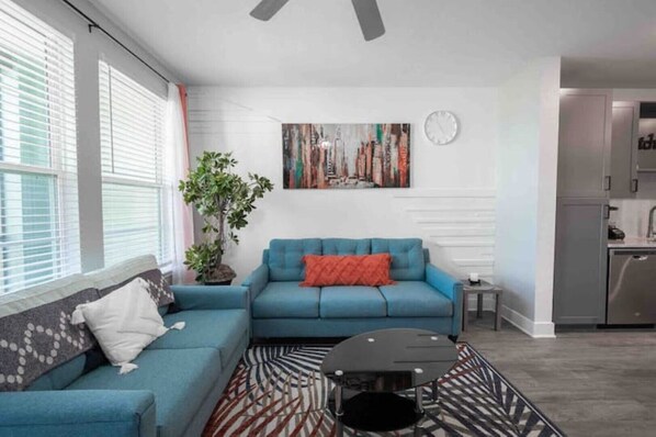 Un salon avec un canapé bleu, un tapis à motifs, une plante en pot et un tableau représentant un paysage urbain accroché au mur.