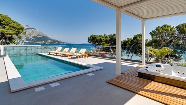 Un espace piscine extérieur moderne, doté de chaises longues et d’un jacuzzi, offrant une vue panoramique sur les montagnes et la mer.