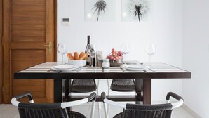 Un coin repas avec une table en bois dressée pour quatre personnes, entourée de chaises en osier. Deux lampes suspendues sont placées au-dessus de la table, et une porte est visible sur la gauche.
