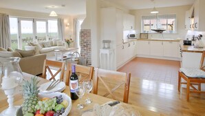 A modern kitchen with white cabinets, a dining area with a wooden table set for a meal, and a living room with a sofa and a view of the outdoors.