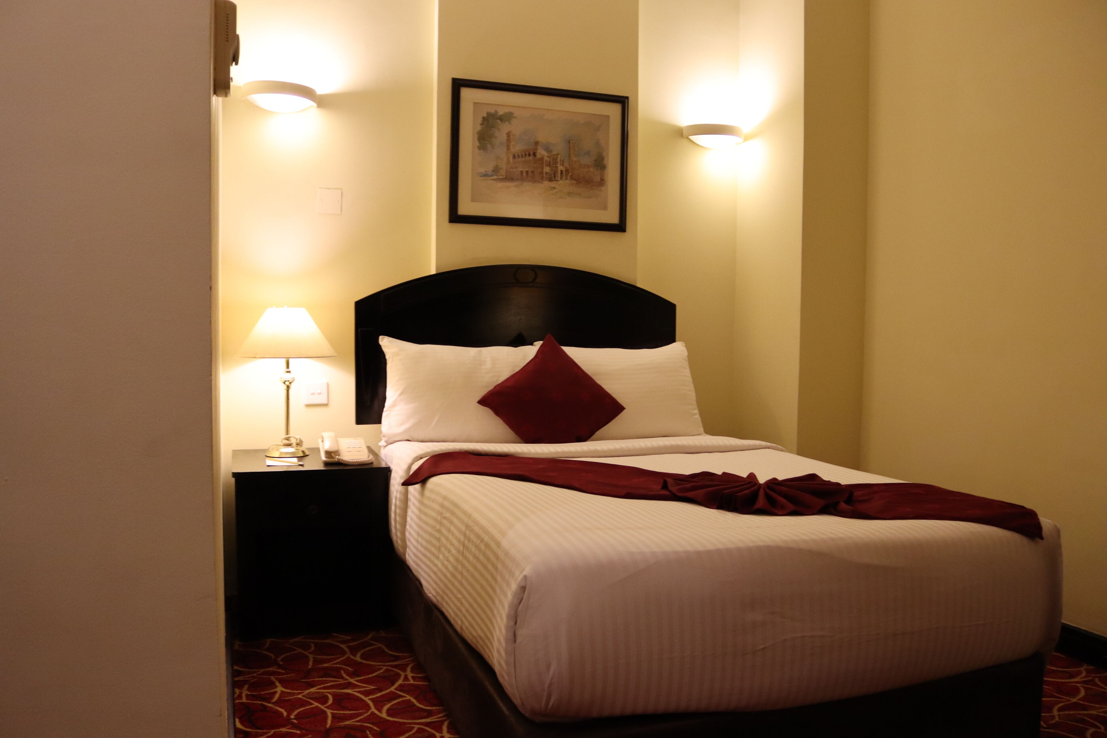 A neatly made bed with a red pillow and a white comforter, a bedside lamp, and a framed picture on the wall.