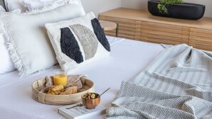 Un lit bien fait, recouvert d’une couverture à motifs blancs et gris, une table de chevet avec un livre et un bol de céréales, et un plateau avec des pâtisseries et du jus de fruits.