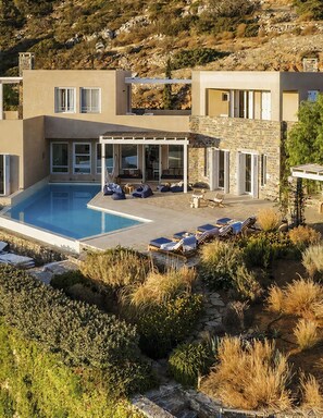 Une maison moderne à deux étages, dotée d’une piscine, d’un espace de détente extérieur et d’une façade en pierre.
