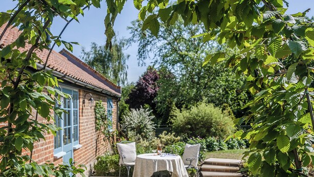 A patio with a table and chairs, a brick building, and surrounding greenery.