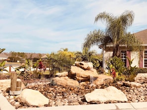 Un jardín ajardinado con un elemento rocoso, palmeras y una casa al fondo.