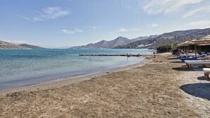Une plage à l’eau claire, avec un rivage rocheux et des montagnes en arrière-plan. On y trouve des chaises longues et une structure en bois sur la droite.