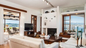 A spacious living room with a fireplace, comfortable seating, a television, and a view of the sea. - Villa Tersanas - Four Bedroom Villa, Sleeps 9 (Livadi)