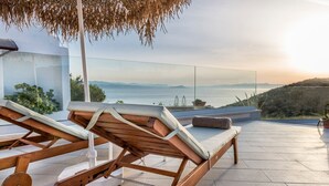 A modern outdoor lounge area with a wooden lounge chair and a bench, overlooking a scenic view of the sea and distant mountains. - Villa Tersanas - Four Bedroom Villa, Sleeps 9 (Livadi)