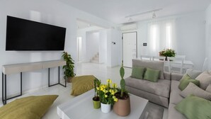 A modern living room with a flat-screen TV, a sofa, a coffee table with plants, and a dining area in the background.