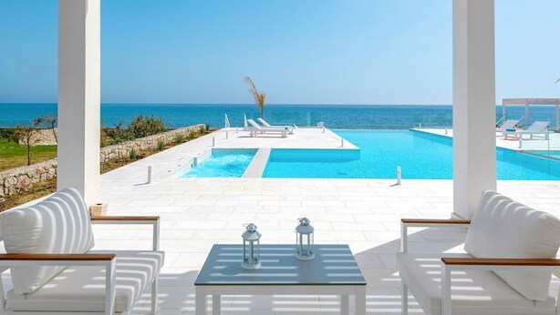 A pool area with white lounge chairs and a table, overlooking a clear blue sea.
