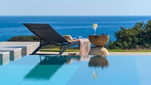 Une chaise longue installée au bord de la piscine, avec un verre de vin et un bol, offrant une vue imprenable sur l’océan bleu azur.