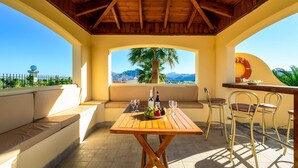 Ein überdachter Außenbereich mit Tisch und Stühlen, Blick auf Berge und eine Palme.