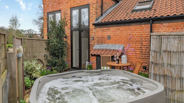 A hot tub in a backyard with a brick house, wooden fence, and a small table with a bottle and glass.
