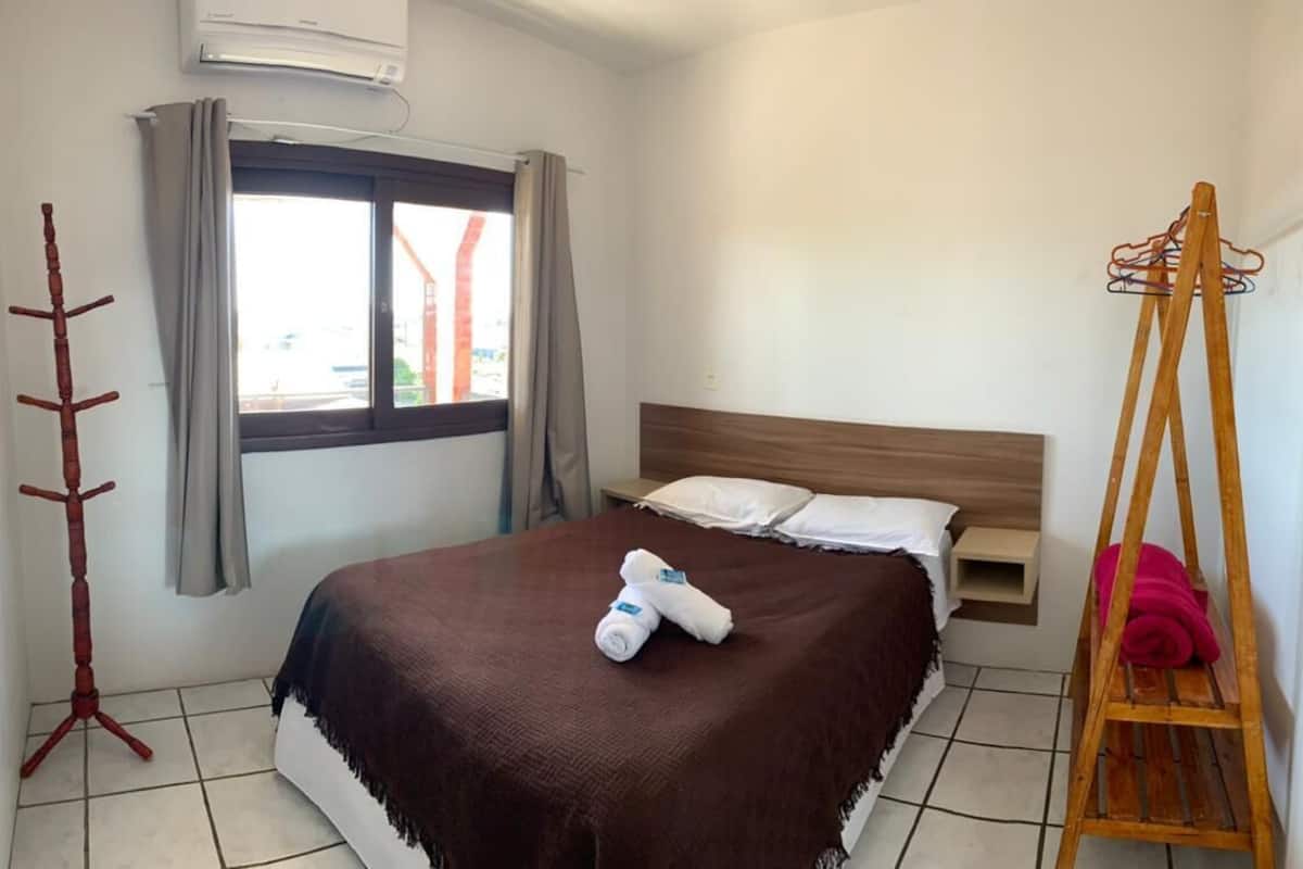 Une chambre à coucher avec un lit, une tête de lit en bois, une table de chevet, une télévision, une fenêtre avec des rideaux, un porte-manteau et une serviette sur le lit.