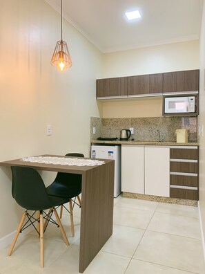 A compact kitchen with a microwave, kettle, and refrigerator. There is a wooden dining table with chairs and a pendant light. - Pousada Dona Monica (Balneario Gaivota)