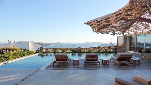A poolside area with lounge chairs, a wooden pergola, and a view of the sea and distant buildings.