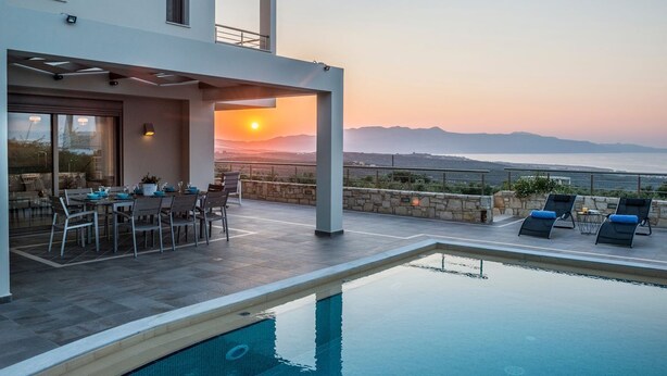 Un área de piscina moderna al aire libre con vistas a la puesta de sol sobre el mar, una zona de comedor con mesas y sillas, y un muro de piedra.