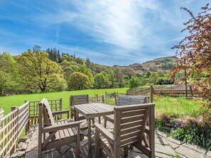 A wooden outdoor seating area with benches and tables, surrounded by a stone wall and a wooden fence, overlooking a green field and hills. - Stybarrow View Cottage (Glenridding, near Ullswater)