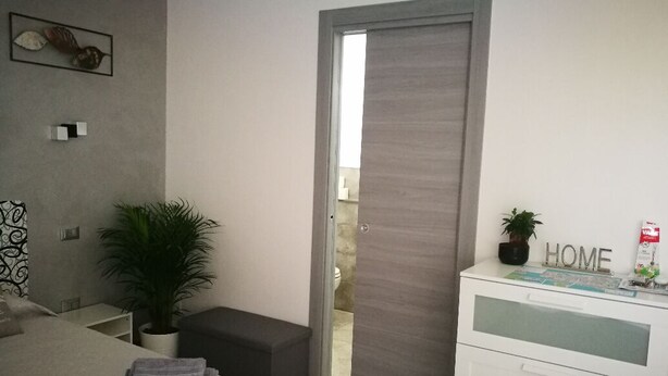 A modern bedroom with a bed, a bedside table, a potted plant, a dresser, and a door leading to a balcony.