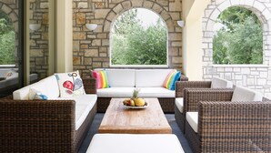 Cómoda zona de estar al aire libre con mobiliario de mimbre, una pared de piedra y ventanas amplias que ofrecen vistas a la vegetación.