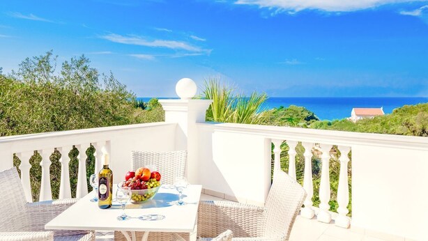 A balcony with wicker furniture, a table with a bottle and fruit, and a view of the sea. - Villa Mellow - Three Bedroom Villa, Sleeps 6 (Ionian Islands)