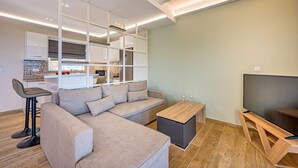 Een moderne woonkamer met een modulaire bank, een houten salontafel, een televisie op een houten dressoir en een keukenhoek met kastjes en een raam.