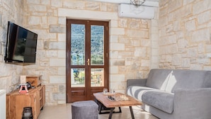 A stone-walled room with a grey sofa, a wooden table with chess pieces, a television mounted on the wall, and a door leading to a balcony. - Villa Peristeri - Six Bedroom Villa, Sleeps 18 (Apokoronos)